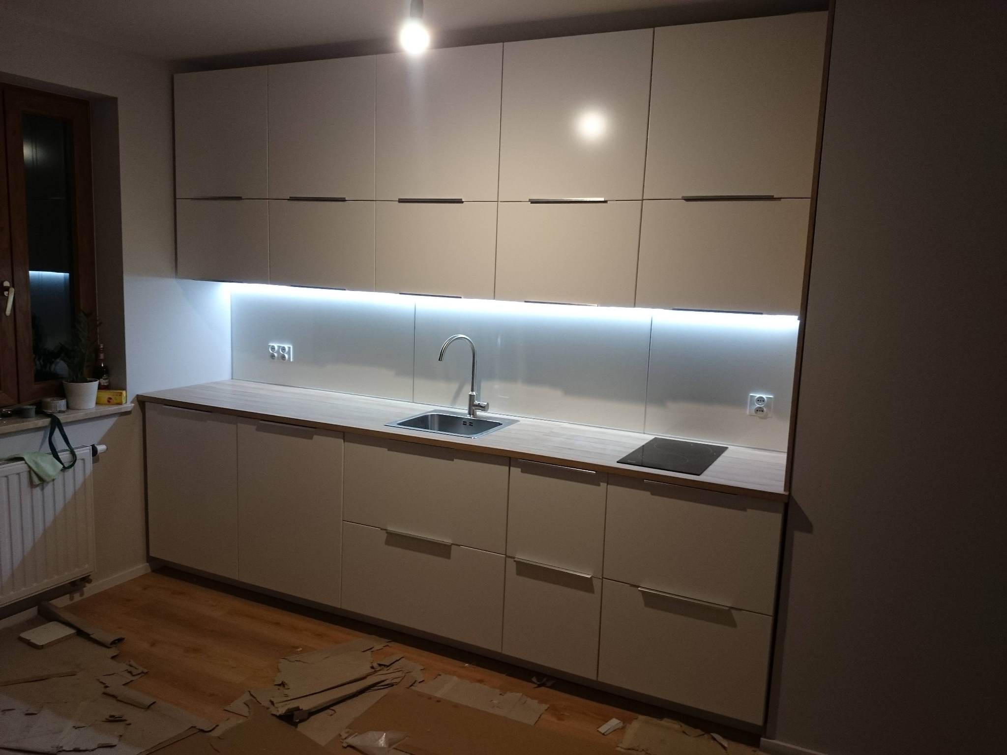 Jasna zabudowa kuchenna z drewnianym blatem, oświetleniem LED pod szafkami wiszącymi, widoczne ślady montażu na podłodze.