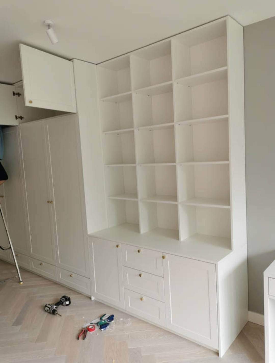 Biała zabudowa meblowa z otwartymi półkami i szafkami z szufladami, widoczne narzędzia na podłodze, wnętrze w trakcie montażu.