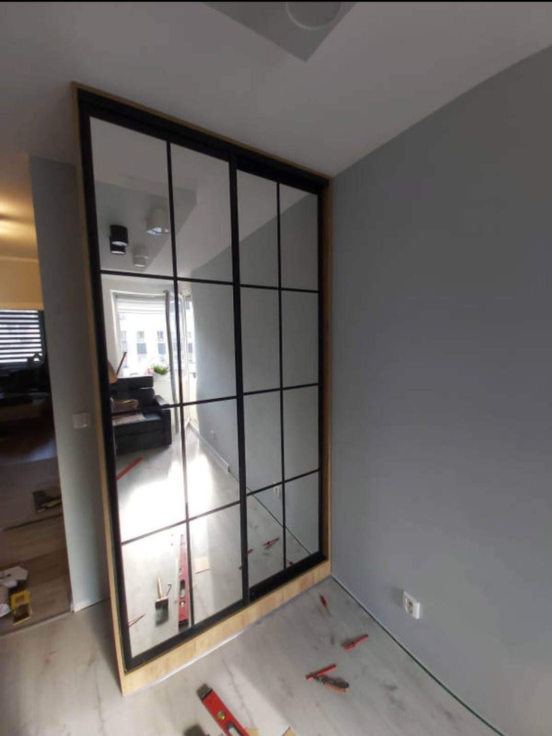 Nowoczesna, trzyskrzydłowa przesuwana zabudowa wnęki z lustrami i czarnymi szprosami, widoczne narzędzia montażowe na podłodze.