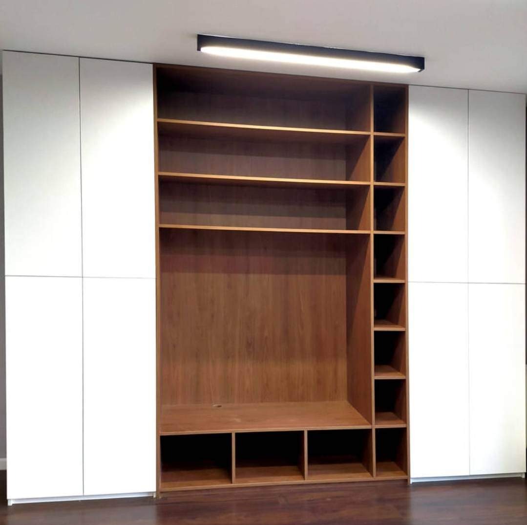 Nowoczesna zabudowa meblowa w Krakowie: białe szafy z frontami bez uchwytów po bokach i otwarta drewniana konstrukcja z półkami i wnękami pośrodku, oświetlona podłużną lampą sufitową.