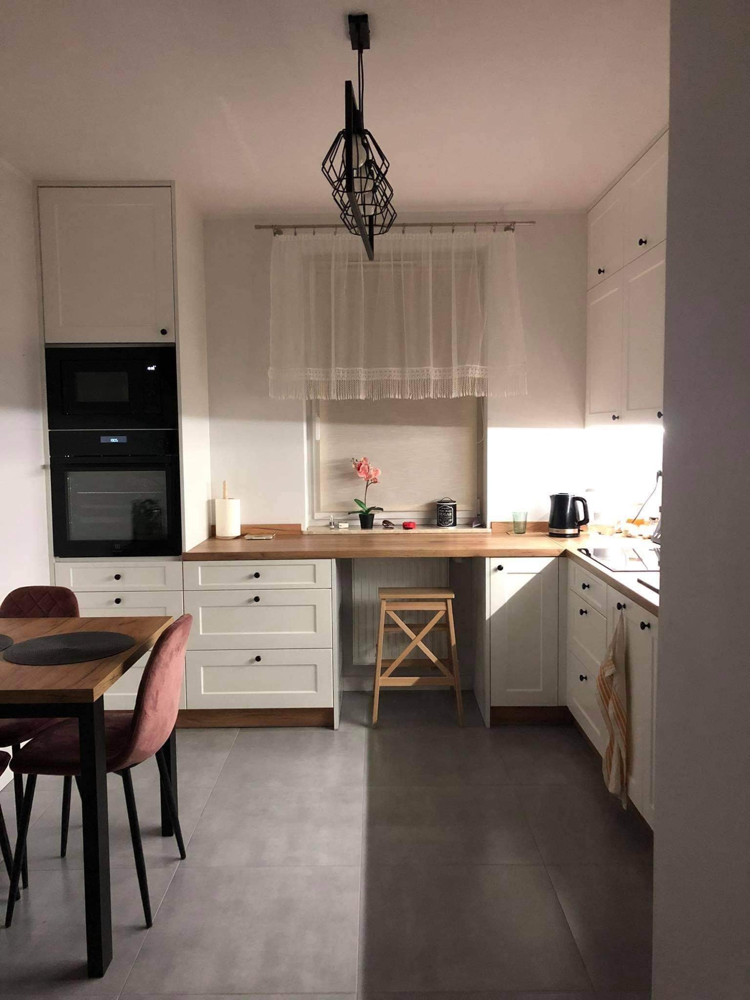 Jasna kuchnia z białymi szafkami, drewnianym blatem, czarnymi uchwytami i nowoczesną lampą wiszącą, stół z aksamitnymi krzesłami w odcieniach różu, drewniany taboret pod oknem.