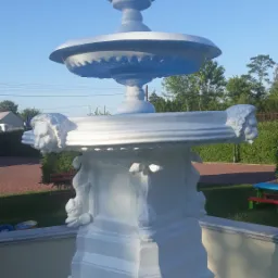 Odnowiona, biała fontanna ogrodowa z motywami lwich głów i ryb, widoczna na tle błękitnego nieba i zieleni drzew.