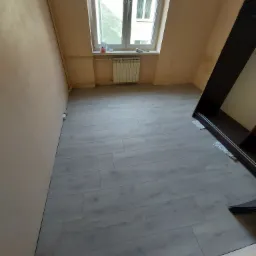 Jasnoszare panele podłogowe ułożone w pokoju z oknem, widoczny kaloryfer pod oknem i fragment szafy z zabezpieczeniami na podłodze.