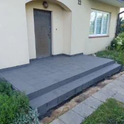 Zewnętrzne schody wejściowe do domu wyłożone szarą, kwadratową terakotą, widoczne ślady prac ziemnych i roślinność.