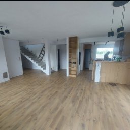 Najmajster Paweł Nowicki - Jasne panele podłogowe w przestronnym wnętrzu domu w trakcie wykańczania, widoczne schody i elementy zabudowy kuchennej. Nowoczesne oświetlenie.