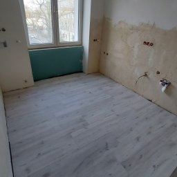Najmajster Paweł Nowicki - Jasne panele podłogowe w pokoju z oknem i przygotowanymi ścianami do wykończenia. Widoczne instalacje elektryczne i hydrauliczne.
