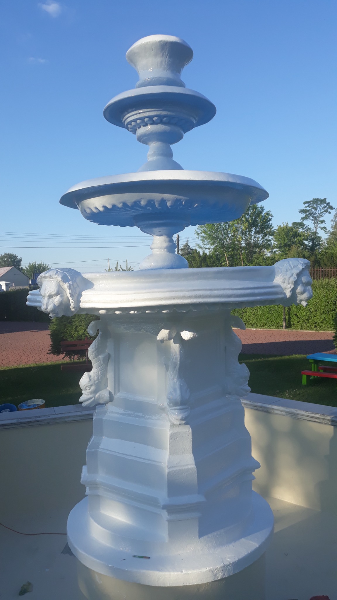 Odnowiona, biała fontanna ogrodowa z motywami lwich głów i ryb, widoczna na tle błękitnego nieba i zieleni drzew.