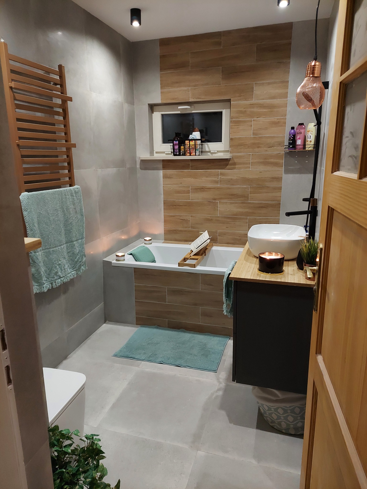 Nowoczesna łazienka z płytkami imitującymi drewno, wiszącą szafką z umywalką nablatową, drewnianym grzejnikiem drabinkowym i dekoracyjnym oświetleniem w postaci miedzianej lampy.