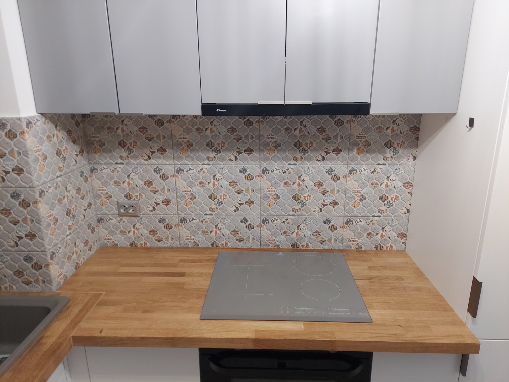 Nowoczesna kuchnia po renowacji: drewniany blat, szara płyta indukcyjna, wzorzyste płytki ścienne i białe szafki wiszące z czarnym okapem.