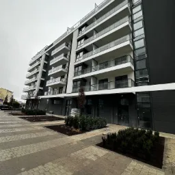 Nowoczesny, wielopiętrowy budynek apartamentowy z balkonami i przeszkloną fasadą, widoczny z poziomu chodnika wyłożonego kostką brukową. Szare niebo w tle.