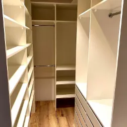 Wnętrze garderoby z drewnianą podłogą i białymi, modułowymi półkami oraz szufladami. Widoczne drążki na ubrania. Nowoczesny design z dbałością o detale.