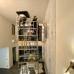 Clean Master 3City Professional cleaning firm - Dwóch mężczyzn na rusztowaniu czyści sufit i ścianę w jasnym wnętrzu. Prace na wysokości, widoczne narzędzia i materiały. Ujęcie z dołu, oświetlenie mieszane.