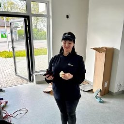 Clean Master 3City Professional cleaning firm - Pracownica firmy sprzątającej w Gdyni, w uniformie, trzymająca ścierkę i telefon, stoi w pustym, wysprzątanym lokalu z otwartymi drzwiami wejściowymi.