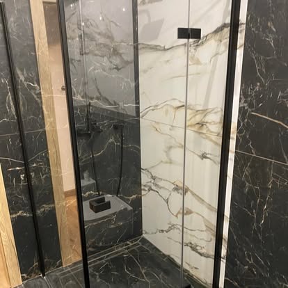 Nowoczesna kabina prysznicowa z czarnymi profilami i marmurowymi płytkami w odcieniach bieli i czerni. Widoczny czarny panel prysznicowy i półka.