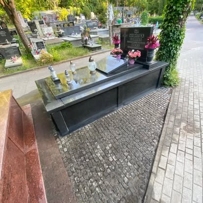 Czarny, wypolerowany nagrobek z kwiatami i lampionami na cmentarzu, widok z boku, kostka brukowa na pierwszym planie, drzewa w tle.