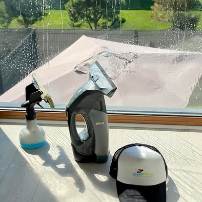 Okno z pianą czyszczącą, spryskiwacz, elektryczna myjka i czapka z logo firmy CleanMaster. Widok na zewnątrz, zieleń i parasol.