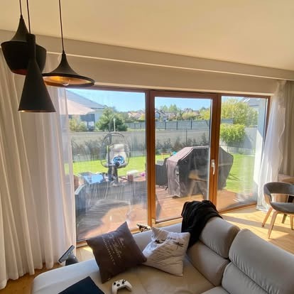 Salon z dużym oknem tarasowym, przez które widać zadbany ogród z huśtawką i grillem. Nowoczesne lampy wiszące i jasna sofa dodają wnętrzu przytulności.