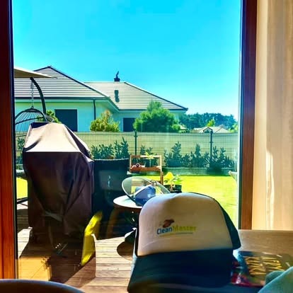 Widok przez okno na zadbany ogród z domu; w pierwszym planie czapka z daszkiem 'Clean Master'.