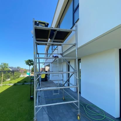 Aluminiowy ruszt na kółkach ustawiony przy białej elewacji nowoczesnego domu z dużymi oknami; widoczne narzędzia i węże. Błękitne niebo w tle.