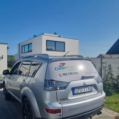 Srebrny samochód z naklejką firmy sprzątającej 'Clean Master' zaparkowany przed nowoczesnym, białym domem w słoneczny dzień. Widoczny numer telefonu.