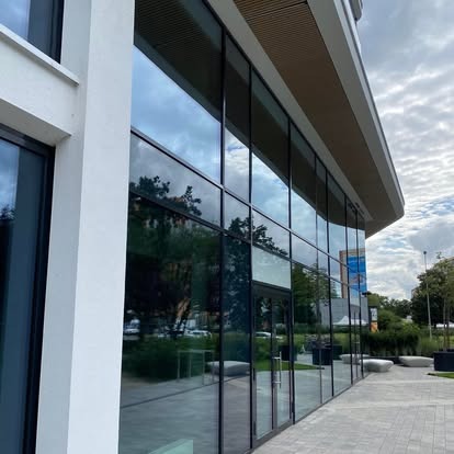 Szklana fasada nowoczesnego budynku biurowego z refleksami nieba i zieleni. Widok z zewnątrz, podkreślający czystość i przejrzystość powierzchni.