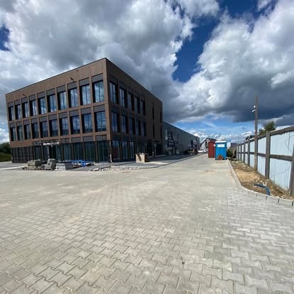 Nowoczesny budynek biurowy z kostką brukową na zewnątrz, widok z poziomu gruntu. Częściowo pochmurne niebo nad obiektem.