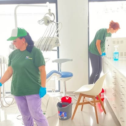 Dwie osoby sprzątają gabinet dentystyczny. Kobieta w zielonej koszulce i fioletowych spodniach oraz druga, pochylona nad szafką. Widoczne krzesło dentystyczne.