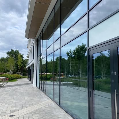 Nowoczesny budynek biurowy z dużymi przeszkleniami, odbijającymi zieleń drzew i błękitne niebo, widok z zewnątrz na chodnik z kostki brukowej i wejście.