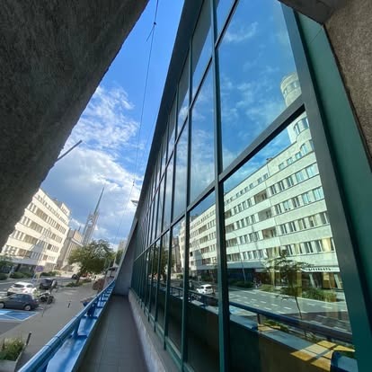 Szklana fasada budynku odbijająca błękitne niebo i architekturę miejską. Widok z boku, ukazujący perspektywę ulicy i okien.