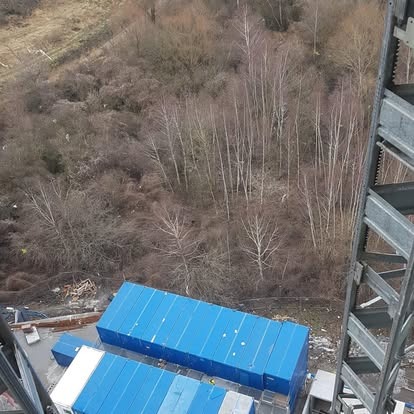 Widok z góry na teren budowy z niebieskimi kontenerami, widoczne drzewa i metalowa konstrukcja. Kąt ujęcia z wysokości.