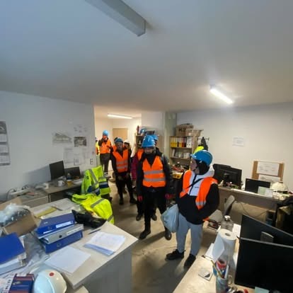 Grupa pracowników w kaskach i kamizelkach odblaskowych w biurze po remoncie, z widocznymi śladami prac budowlanych na biurkach i podłodze. Przygotowania do sprzątania.