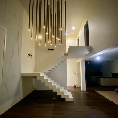 Nowoczesne wnętrze domu z białymi schodami, wiszącą lampą z rurek i ciemną podłogą. Minimalistyczny design z oświetleniem akcentującym.