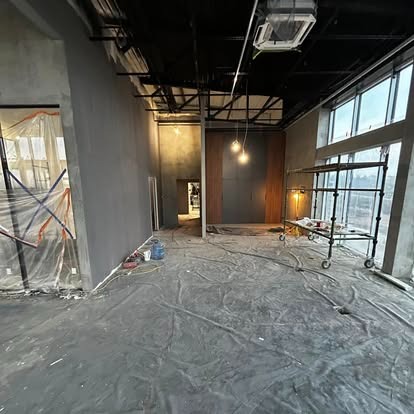 Wnętrze w trakcie wykańczania: szpachlowane ściany, zabezpieczona folią podłoga, rusztowanie przy oknie, widoczne instalacje pod sufitem i wiszące lampy.