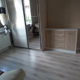 Jasne panele podłogowe w pokoju z beżowymi ścianami, widoczna szafa z lustrem i komoda z szufladami.