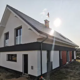 Nowoczesny dom jednorodzinny z białą elewacją i ciemnymi akcentami, widoczny fragment dachu z ciemnej dachówki, na tle błękitnego nieba z przelatującym ptakiem, ujęcie z niskiej perspektywy.