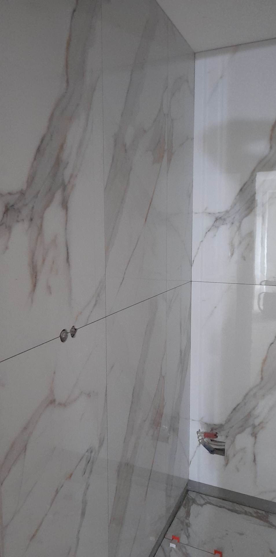 Ściana i narożnik wyłożone białymi płytkami imitującymi marmur, widoczne otwory na gniazdka elektryczne i fragment instalacji hydraulicznej, na podłodze krzyżyki dystansowe.