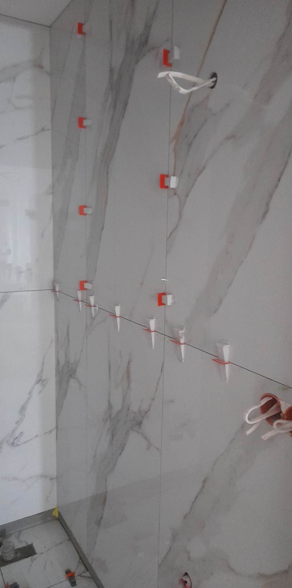 Ściana z marmurowymi płytkami w trakcie montażu, z systemem poziomowania płytek (klipsy i kliny) oraz sznurkiem wyznaczającym linię, widoczne przewody elektryczne wystające ze ściany.