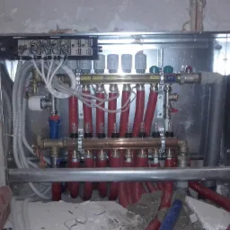 Rozdzielacz ogrzewania podłogowego w metalowej szafce, widoczne rury czerwone, zawory termostatyczne i okablowanie, w tle surowa ściana w trakcie remontu.