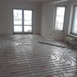 Białe rury ogrzewania podłogowego ułożone na folii z czerwoną siatką w surowym, jasnym wnętrzu z dużymi oknami wychodzącymi na zimowy krajobraz.