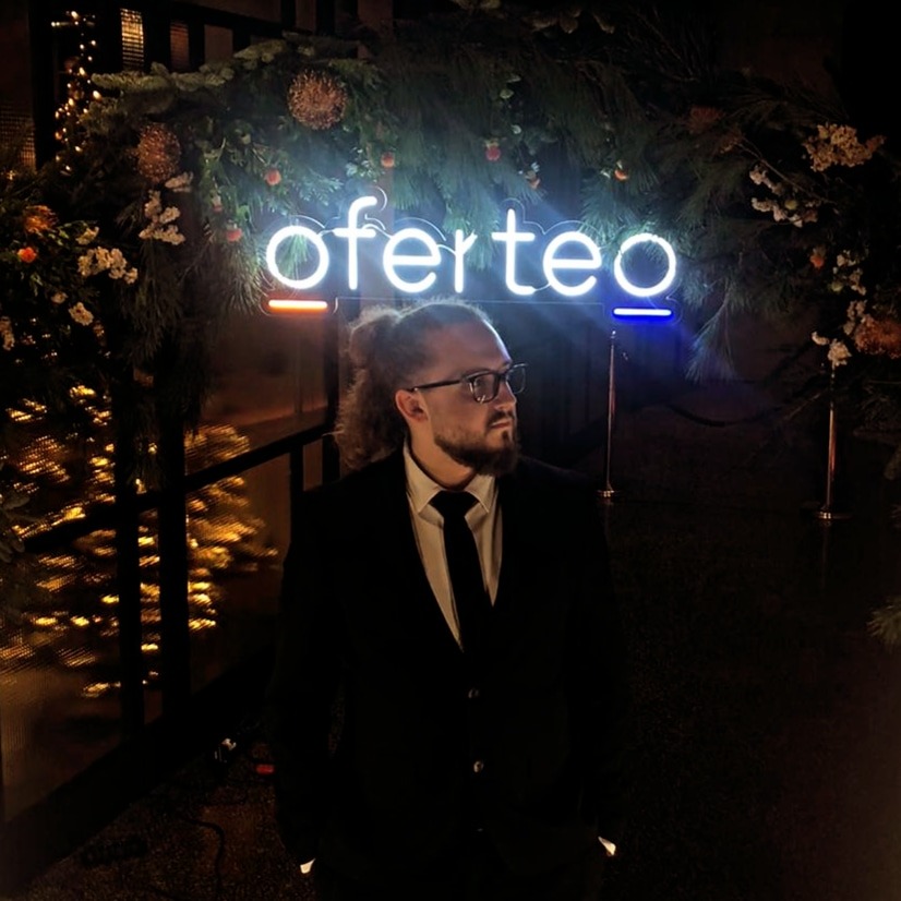 Mężczyzna w garniturze stoi przed neonem 'oferteo' otoczonym dekoracjami roślinnymi i złotymi ozdobami.