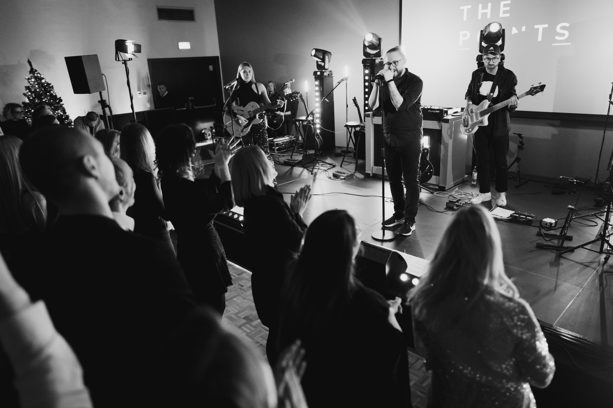 Czarno-białe zdjęcie zespołu muzycznego występującego na scenie przed publicznością, z wokalistą w centrum, gitarzystką po lewej stronie w błyszczących spodniach i gitarzystą po prawej...