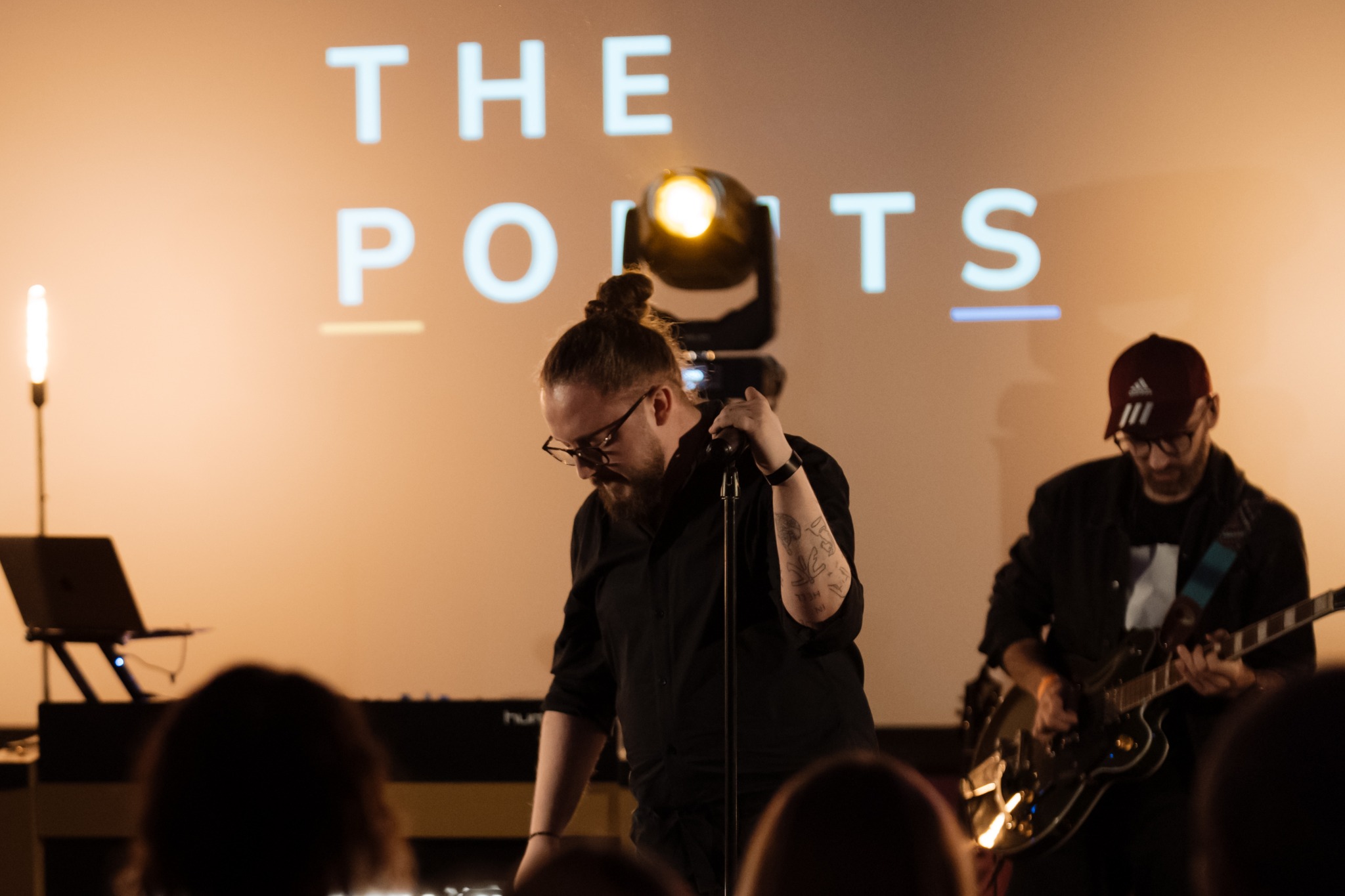 Wokalista z tatuażem na ramieniu trzyma mikrofon na scenie, w tle gitarzysta i wyświetlany napis 'THE POINTS'.