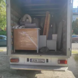 Wnętrze busa transportowego załadowane meblami i przedmiotami, zabezpieczonymi folią stretch, widoczny numer rejestracyjny pojazdu GKW 1XG7