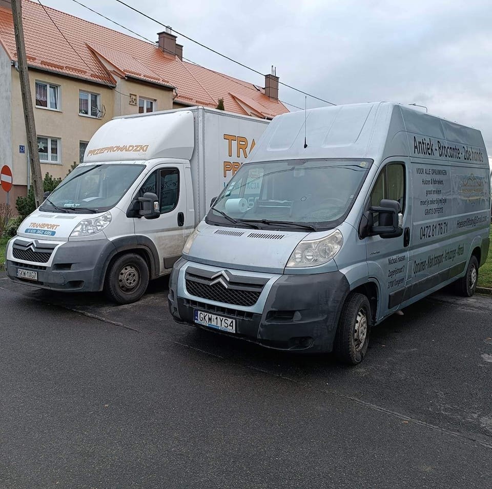 Dwa busy Citroen zaparkowane na ulicy, jeden biały z napisem 'Przeprowadzki' i numerem telefonu, drugi szary z holenderskim napisem 'Antiek-Brocante-2dehands' i numerem telefonu, na tle budynków...