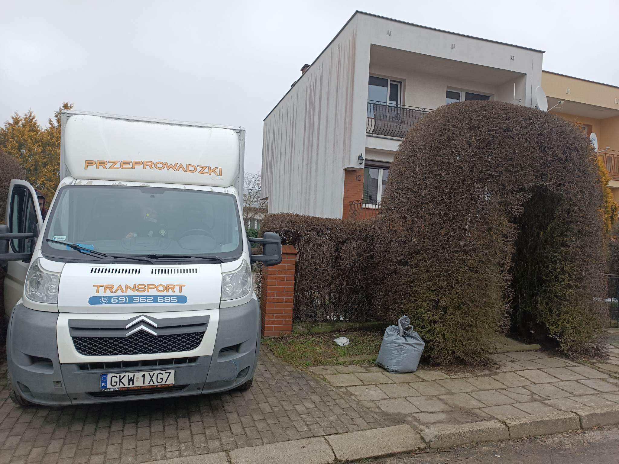 Biały samochód dostawczy z napisem 'PRZEPROWADZKI' i numerem telefonu zaparkowany przed domem z oryginalnie przyciętym żywopłotem w kształcie łuku.