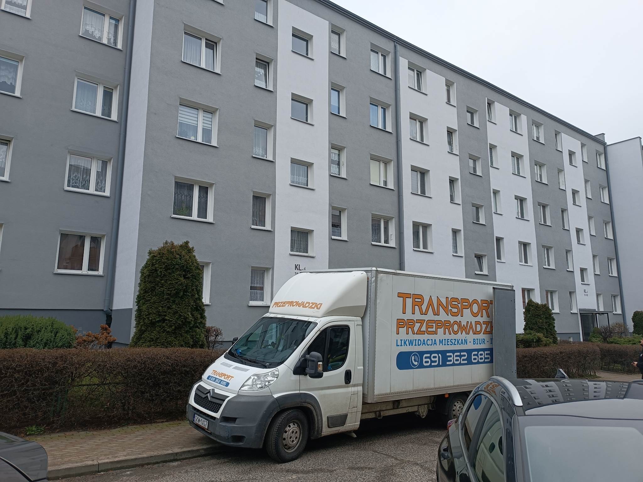 Biały samochód dostawczy z pomarańczowym napisem na tle szarego bloku mieszkalnego, częściowo zasłonięty przez zaparkowany samochód osobowy.