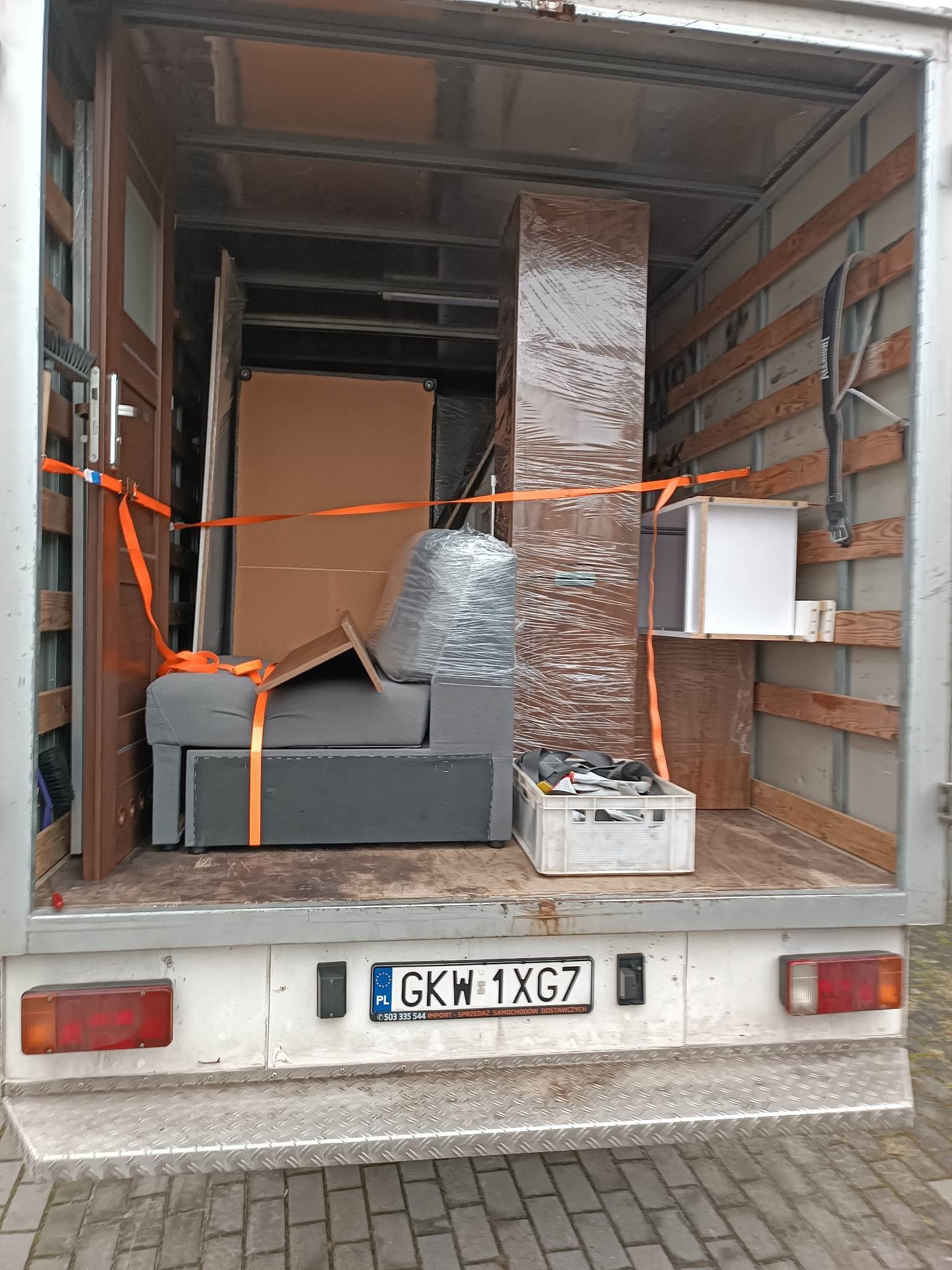 Wnętrze furgonetki załadowane meblami i kartonami, zabezpieczone pomarańczowymi pasami transportowymi, widoczna tablica rejestracyjna GKW 1XG7.