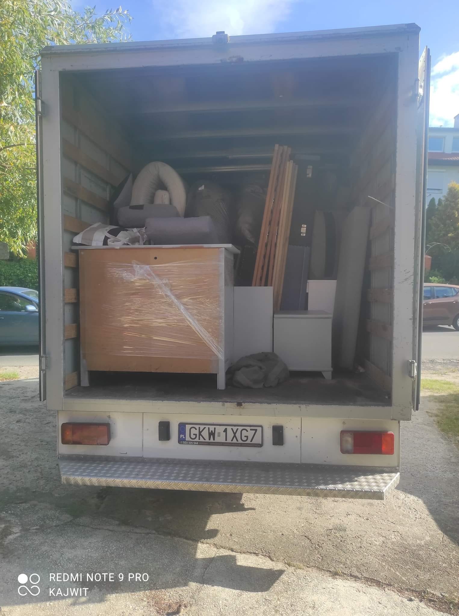 Wnętrze busa transportowego załadowane meblami i przedmiotami, zabezpieczonymi folią stretch, widoczny numer rejestracyjny pojazdu GKW 1XG7