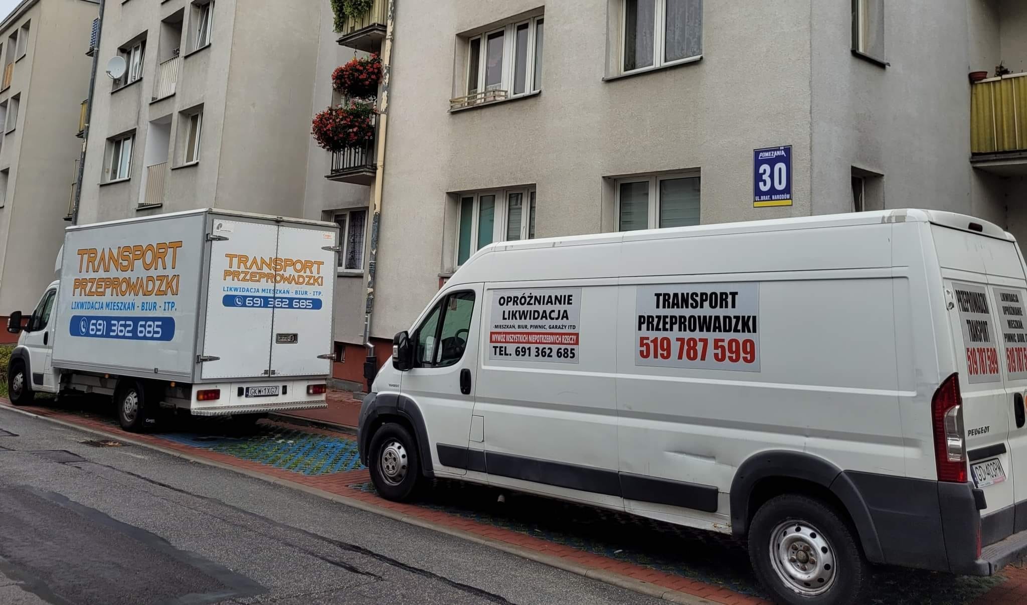 Dwa białe busy transportowe zaparkowane przed budynkiem mieszkalnym z balkonami, z widocznymi numerami telefonów i nazwą firmy na karoserii.