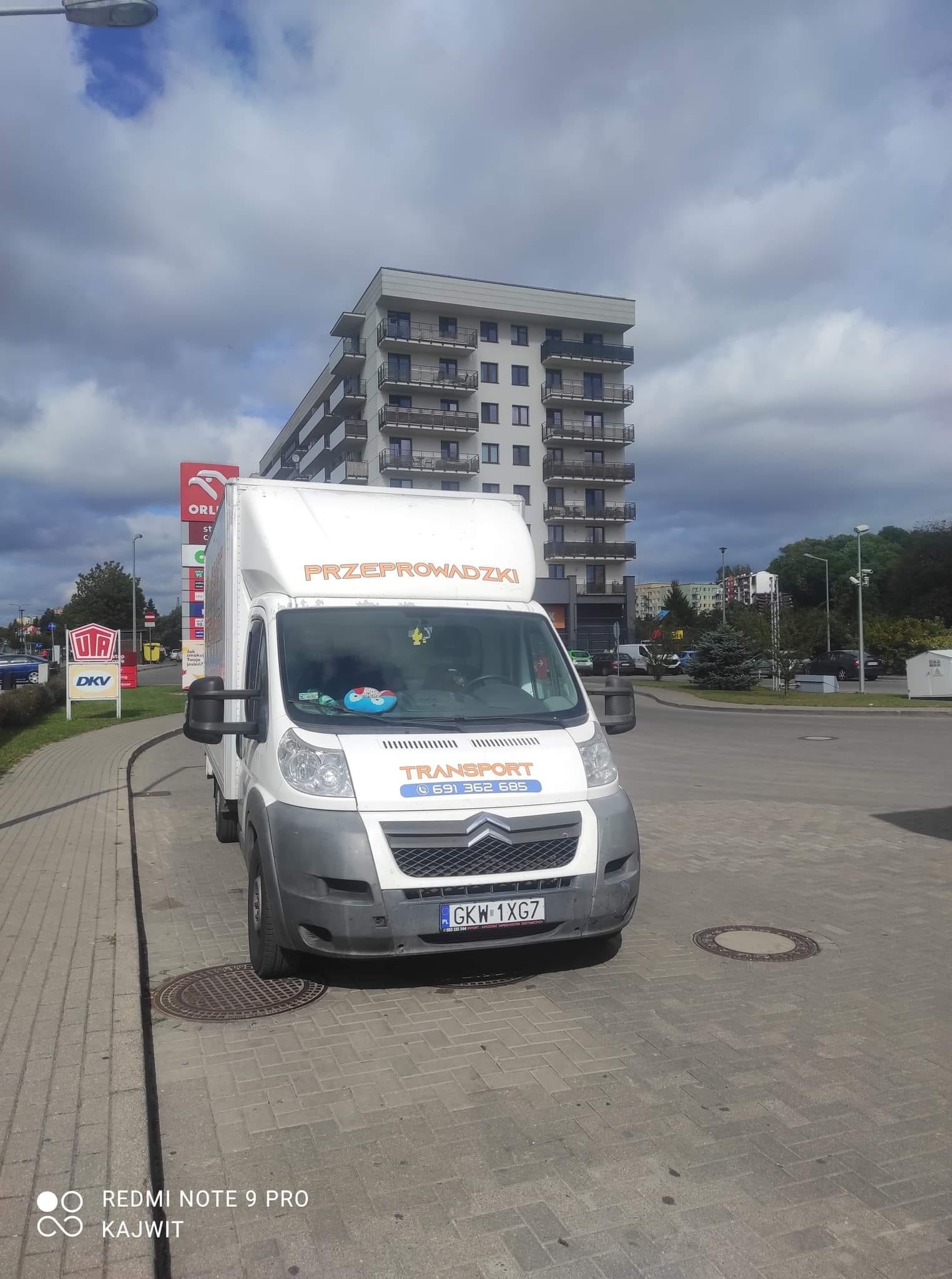 Biały samochód dostawczy z napisem 'PRZEPROWADZKI' zaparkowany na chodniku obok stacji paliw, w tle nowoczesny budynek mieszkalny z balkonami.